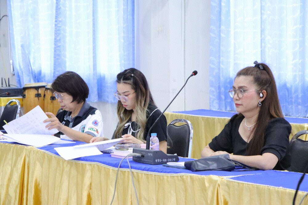 ผู้หญิงสามคนสวมแว่นตานั่งประชุมที่โต๊ะยาว มีเอกสารและไมโครโฟนตั้งอยู่ด้านหน้า