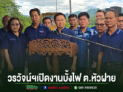กลุ่มคนสวมเสื้อสีน้ำเงินร่วมพิธีเปิดงานบั้งไฟ ต.หัวฝาย โดยมีชายคนหนึ่งยืนพูดที่โพเดียมไม้แกะสลัก