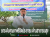 ชายสวมเสื้อสีขาวพูดใส่ไมโครโฟน หน้าป้ายโครงการด้านสาธารณสุขแก่ประชาชน