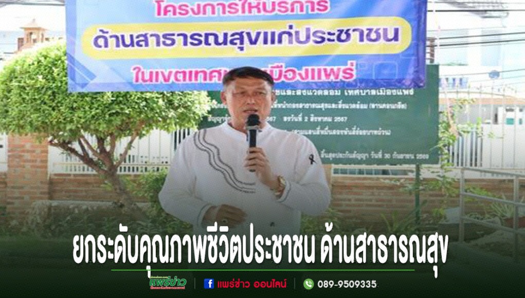 ชายสวมเสื้อสีขาวพูดใส่ไมโครโฟน หน้าป้ายโครงการด้านสาธารณสุขแก่ประชาชน