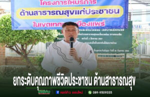 ชายสวมเสื้อสีขาวพูดใส่ไมโครโฟน หน้าป้ายโครงการด้านสาธารณสุขแก่ประชาชน