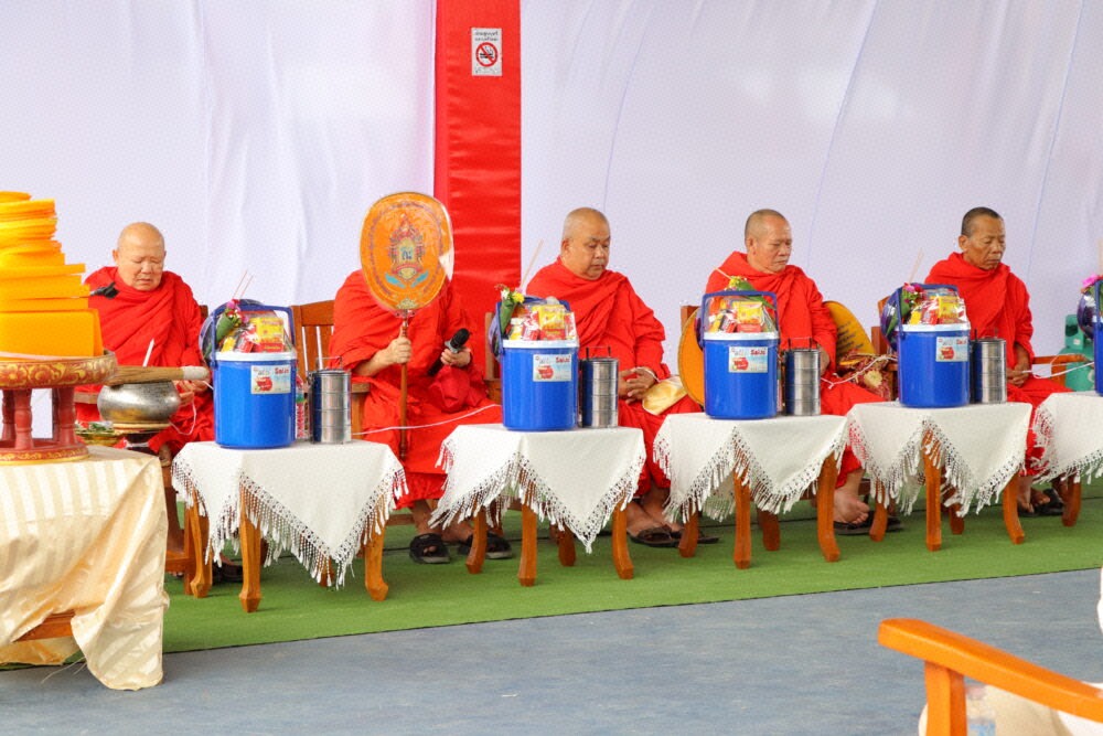 พระสงฆ์ 5 รูปในจีวรสีแดงเข้มนั่งเรียงกันบนเวที มีถังน้ำสีน้ำเงินและปิ่นโตวางอยู่บนโต๊ะเล็กด้านหน้าของแต่ละรูป