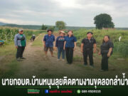 กลุ่มคนยืนถ่ายรูปบนถนนลูกรังกลางทุ่งนา มีข้อความภาษาไทยด้านล่างภาพ