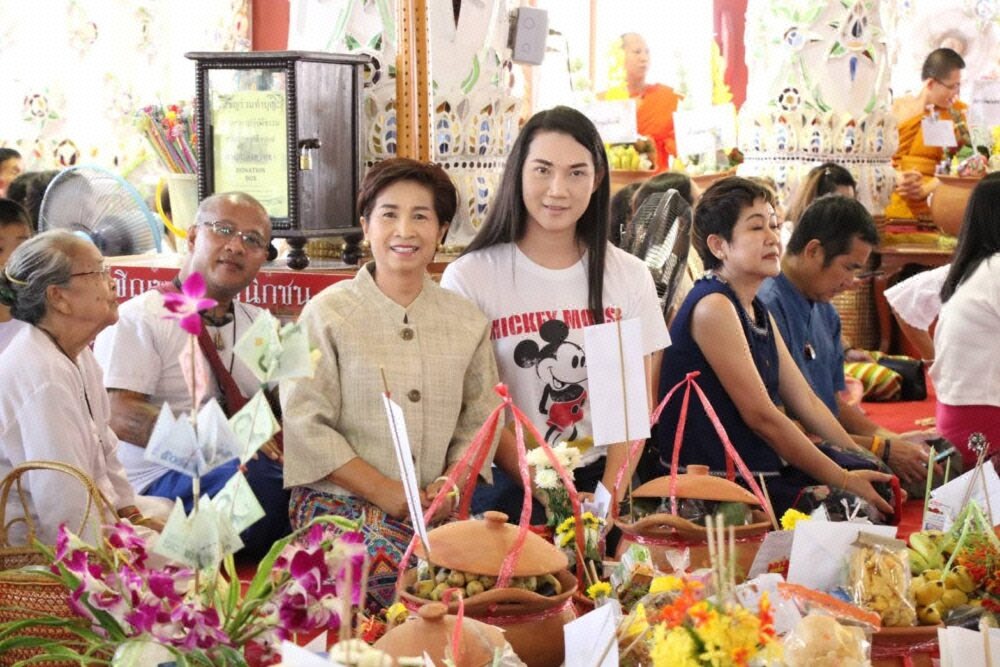 กลุ่มคนนั่งร่วมพิธีทำบุญในวัด มีการจัดเตรียมเครื่องถวายทานและพุ่มเงินตั้งอยู่ด้านหน้า