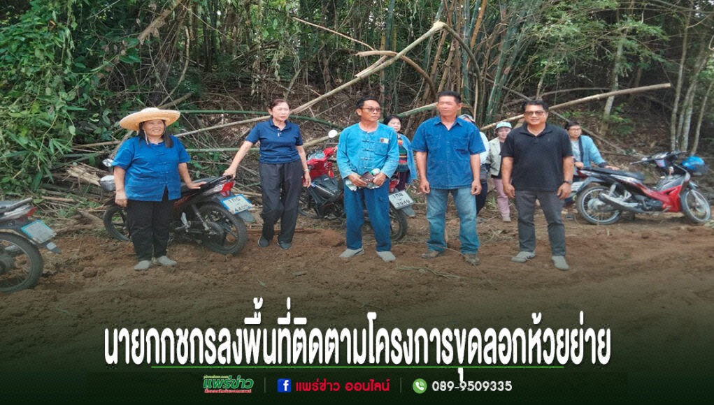 กลุ่มคนยืนถ่ายรูปบนถนนดินลูกรัง มีป้ายข้อความภาษาไทยด้านล่างภาพ