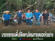 กลุ่มคนยืนถ่ายรูปบนถนนดินลูกรัง มีป้ายข้อความภาษาไทยด้านล่างภาพ