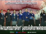 กลุ่มคนยืนเรียงแถวหน้าเวทีที่มีป้ายภาษาไทยและภาษาอังกฤษ พร้อมข้อความภาษาไทยด้านล่างภาพ